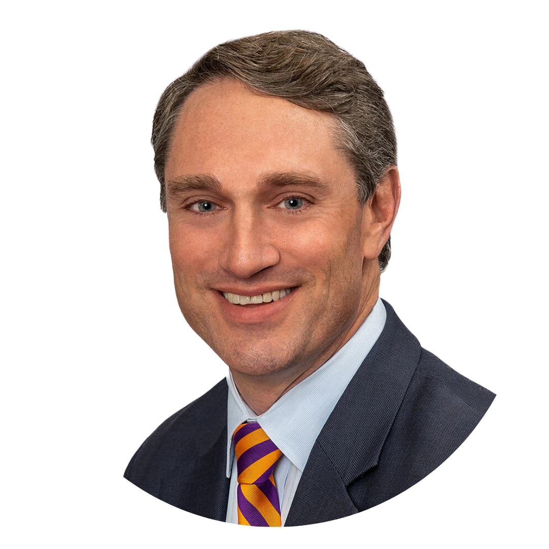 A professional headshot of a man with light brown hair, wearing a dark suit and blue shirt, smiling at the camera. The background is plain white, and the photo is cropped in a circular shape.
