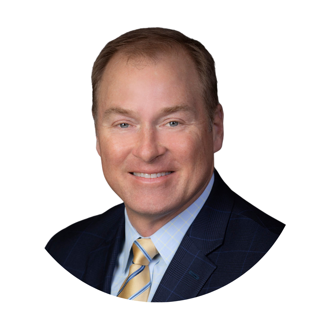 A man wearing a navy blue suit with a light blue shirt and a striped tie, smiling against a plain white background. His hair is short and light brown, and he has a rounded face.
