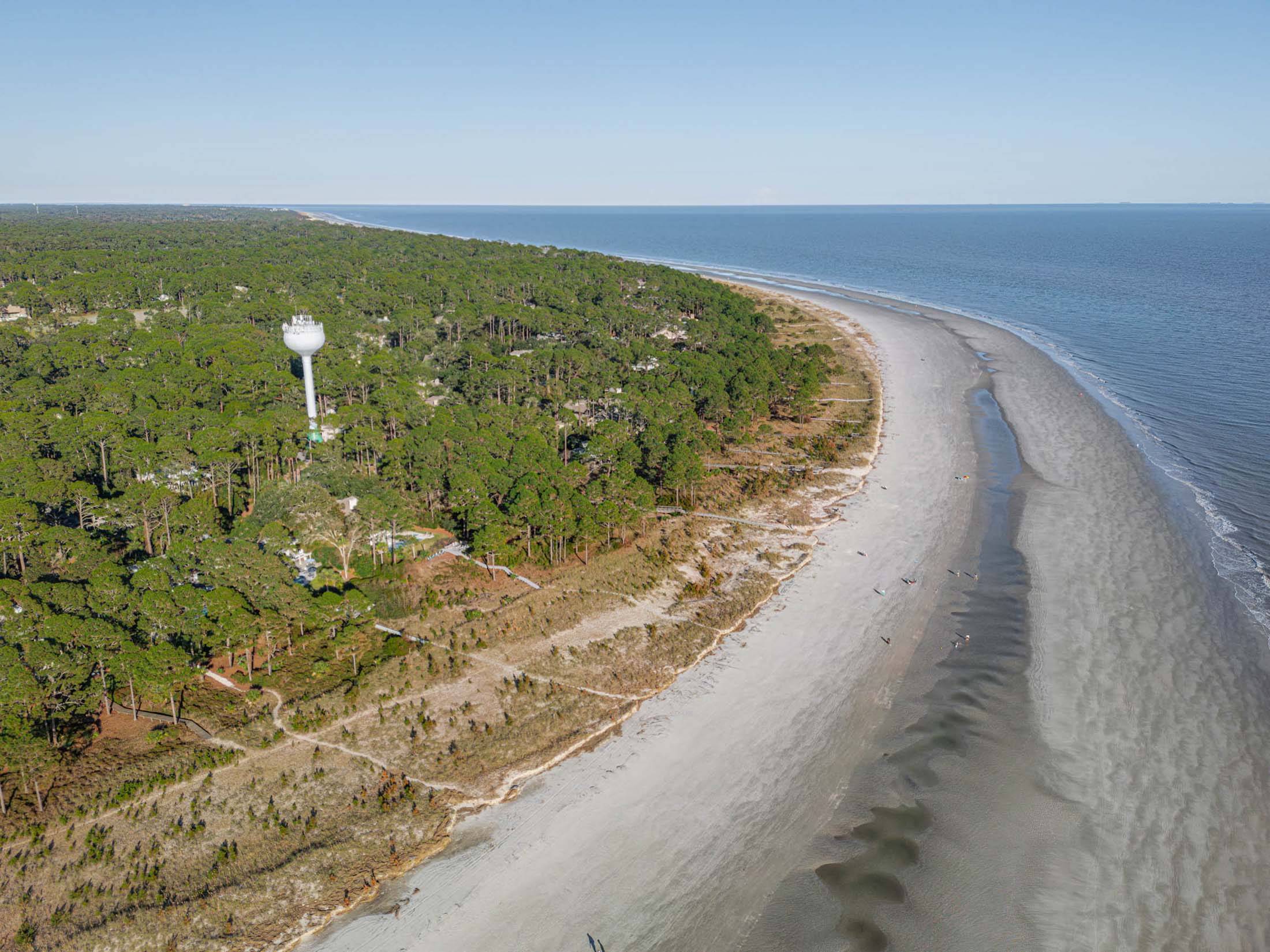 Aerial view of a sandy beach curving along a coastline, bordered by lush green trees. A water tower is visible among the trees, with calm blue ocean water in the distance under a clear sky.