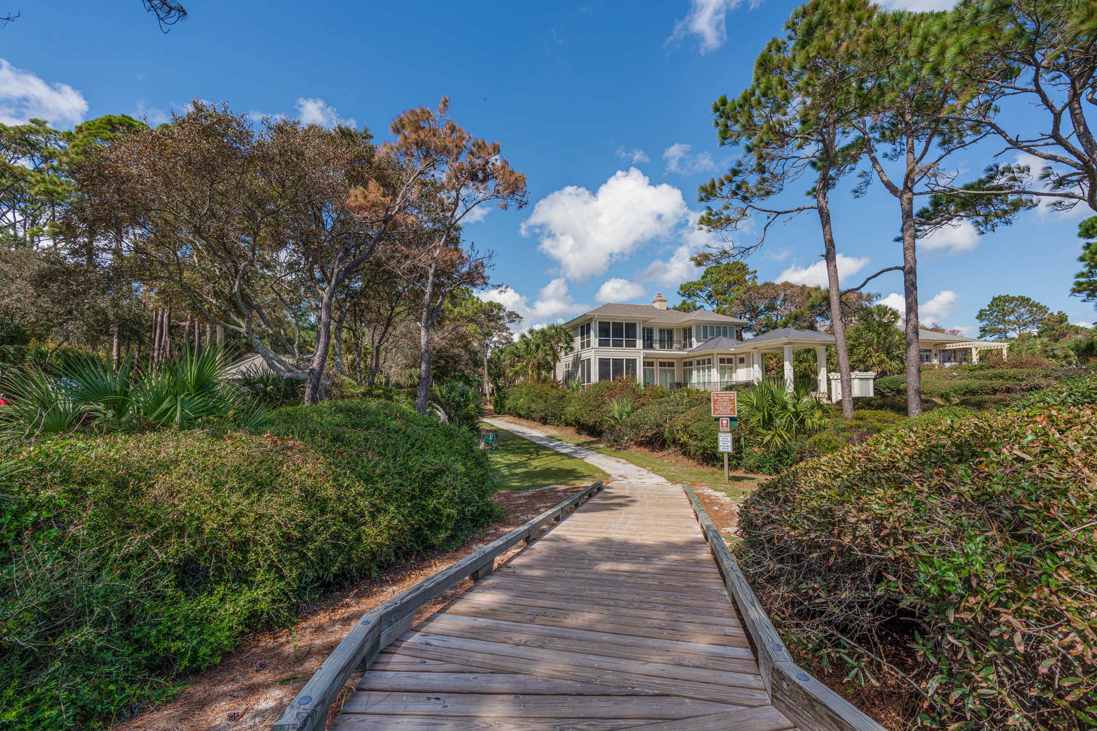 A wooden boardwalk surrounded by lush green bushes and trees, leading to a large house in the background. The sky is bright with white clouds.