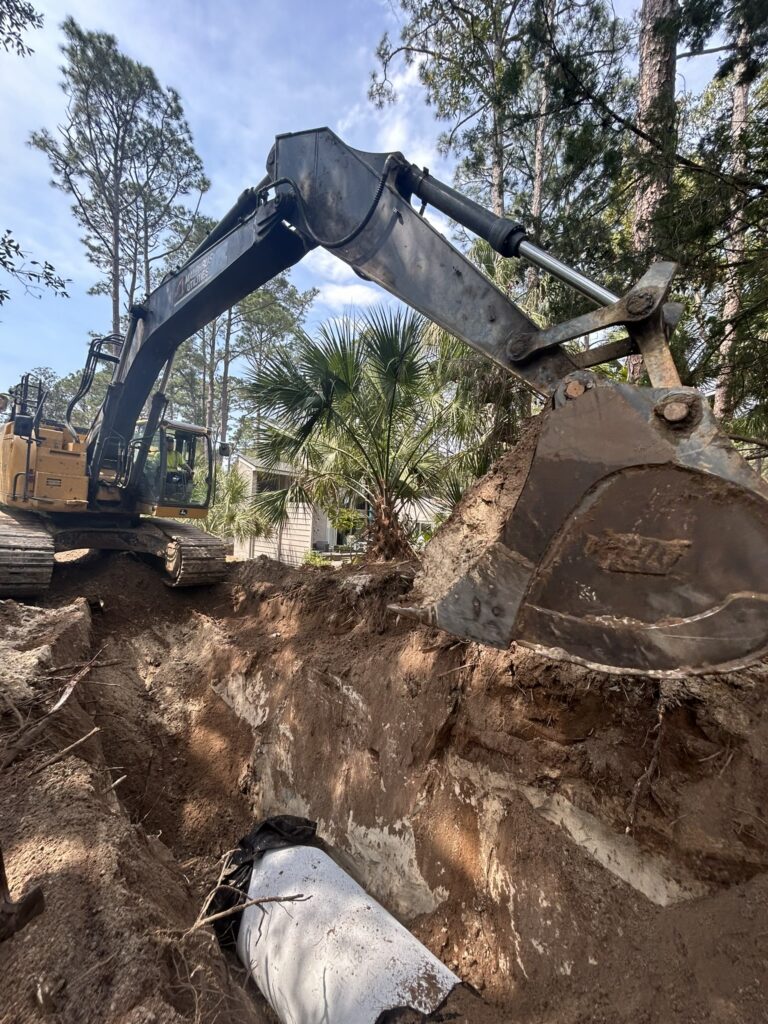 An excavator digging a trench in a wooded area, with a large bucket hovering above the earth. The scene shows a mixture of dirt and vegetation, alongside a visible pipe in the trench.