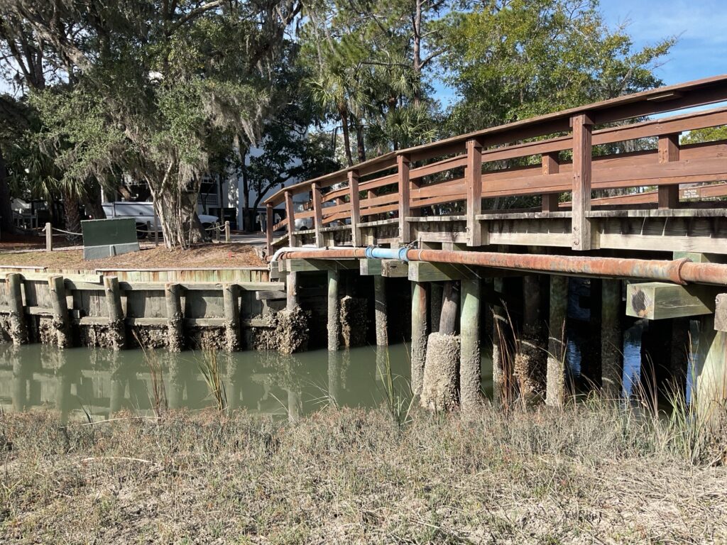 A wooden boardwalk with a railing extends over a marshy area, leading to a dock supported by wooden pilings. The water is shallow and murky, with patches of grass visible along the banks and a backdrop of trees.