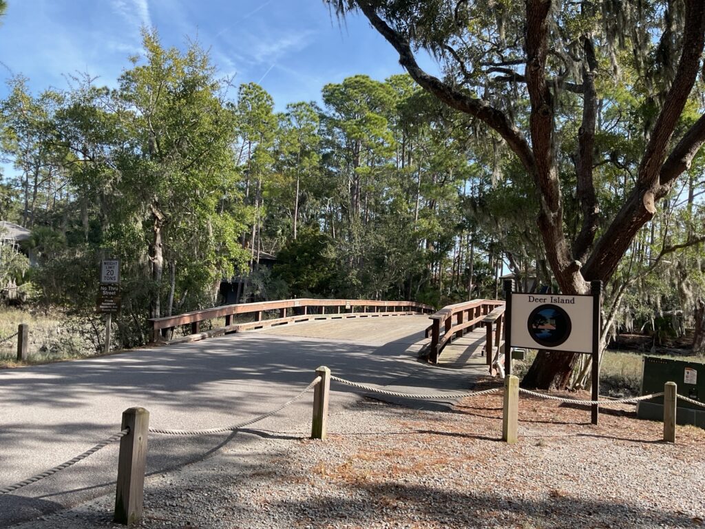 A wooden bridge leading into a wooded area with trees and Spanish moss. A sign for "Deer Island" is visible on the right side of the road, marking the entrance. The scene is sunny with a clear blue sky.
