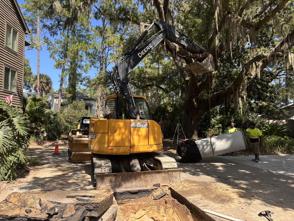 A yellow John Deere excavator removes a large tree trunk on a residential street lined with tall trees and workers in safety vests.