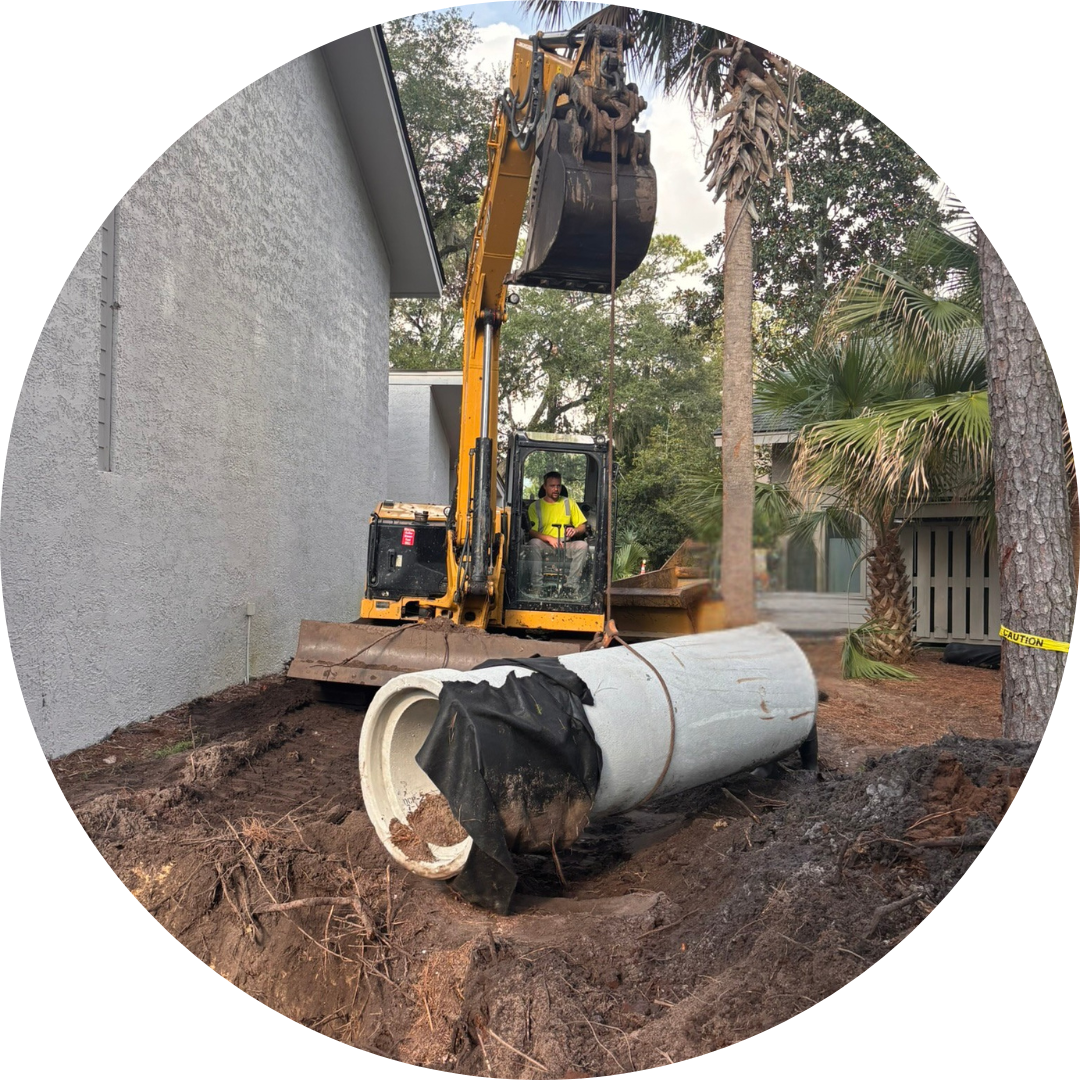 A yellow excavator with an operator inside lifts soil beside a gray building, with a large concrete pipe in the foreground and palm trees nearby.