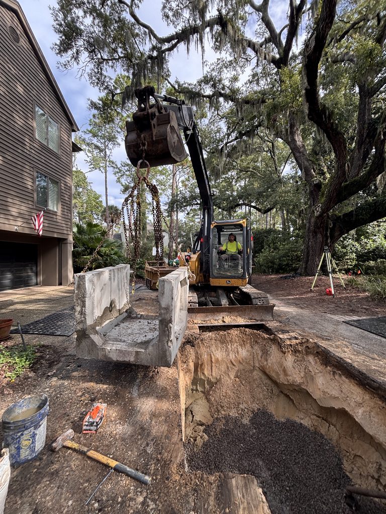 A compact excavator works beside a house foundation on a wooded construction site, with concrete forms, tools, and a dirt path around the area.