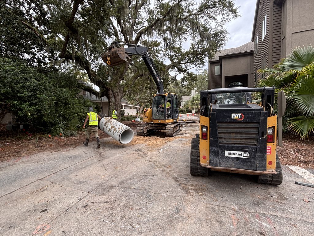 An excavator lifts a large concrete pipe while a worker in a safety vest walks beside it, with a compact CAT loader parked on a residential street.