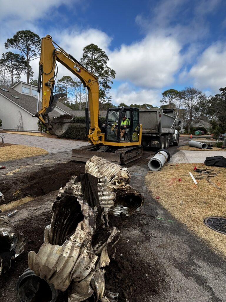 A construction site showing a yellow excavator digging near a house, with a nearby dump truck parked. Large, damaged pipes are visible on the ground, and there is a grassy area with some tools and a utility cover nearby under a partly cloudy sky.
