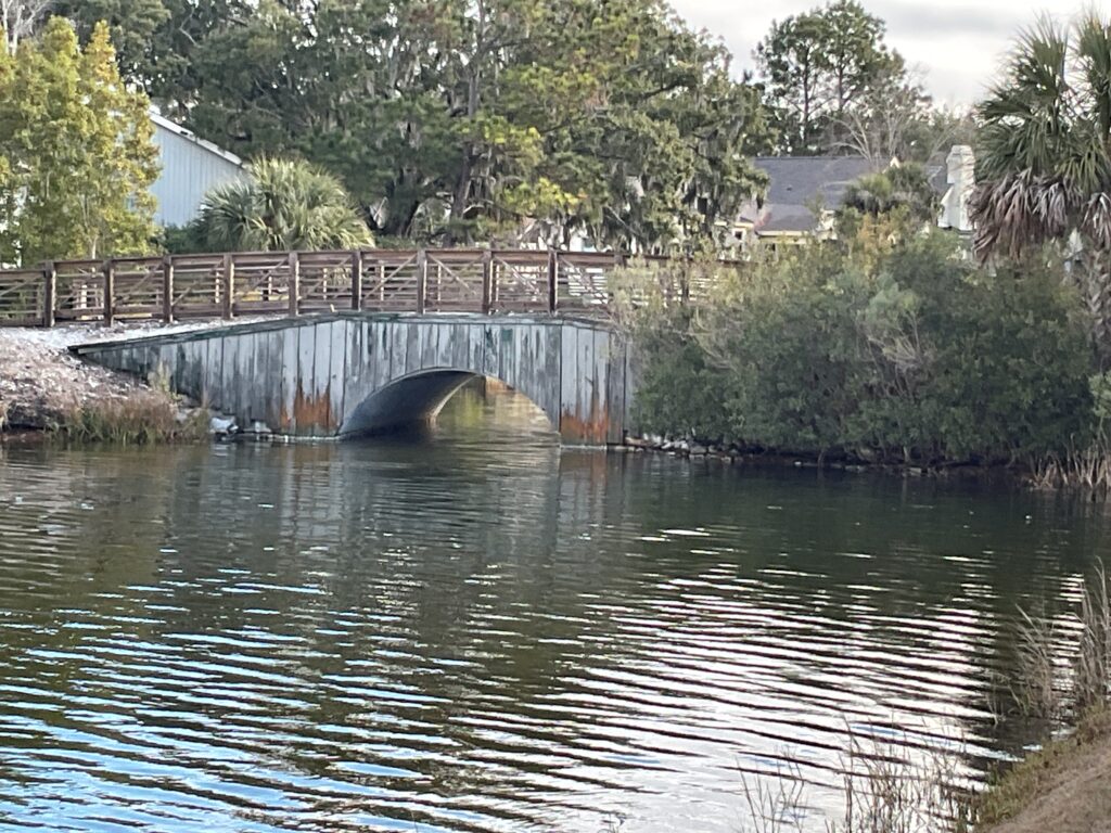 A wooden bridge with a curved arch spans over a calm body of water, reflecting the surrounding greenery. Lush trees and shrubs line the banks, while a few buildings are visible in the background, under a partly cloudy sky.