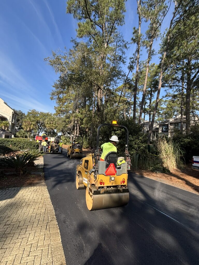Construction workers operating rollers on a newly paved road surrounded by trees in a residential area. The view shows multiple rollers lined up, with workers wearing safety gear, and a blue sky overhead.
