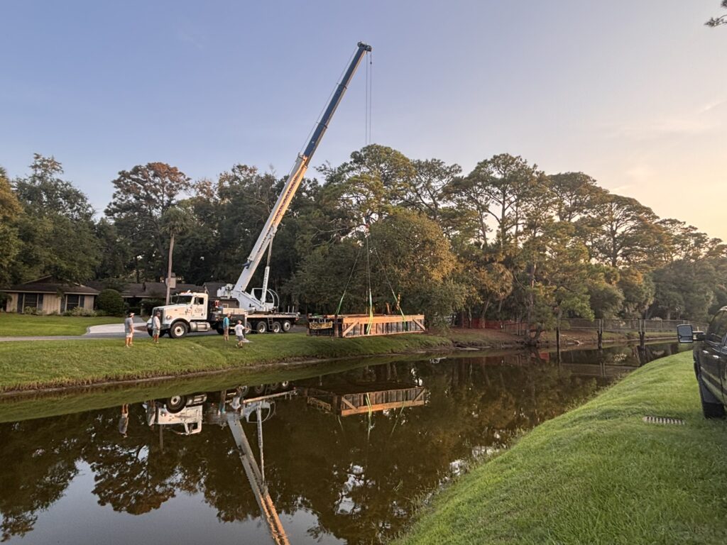 A crane truck with an extended boom stands beside a canal or pond, with workers and a flatbed trailer on the grassy bank at sunset.