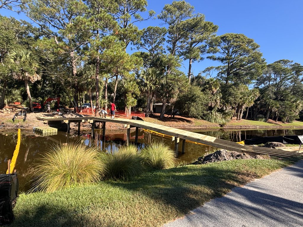 A wooden pedestrian bridge crosses a small pond in a park, surrounded by tall trees, grass, and a paved path.