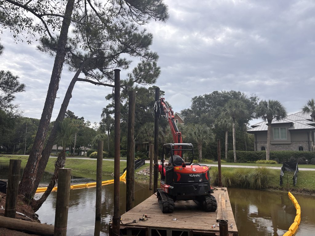 A red Kubota compact excavator sits on a wooden dock beside a canal, with tall vertical posts and trees under a cloudy sky.