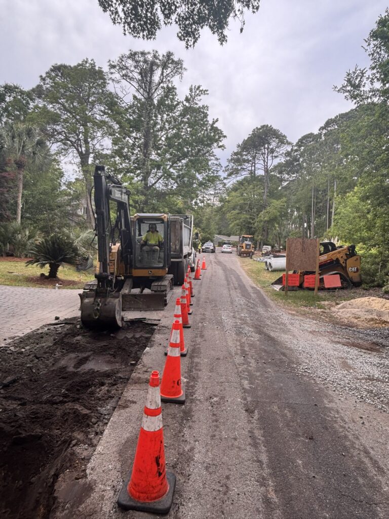 A construction site on a narrow road lined with orange traffic cones. An excavator is positioned near the edge of an open trench, surrounded by greenery and several construction vehicles visible in the distance.